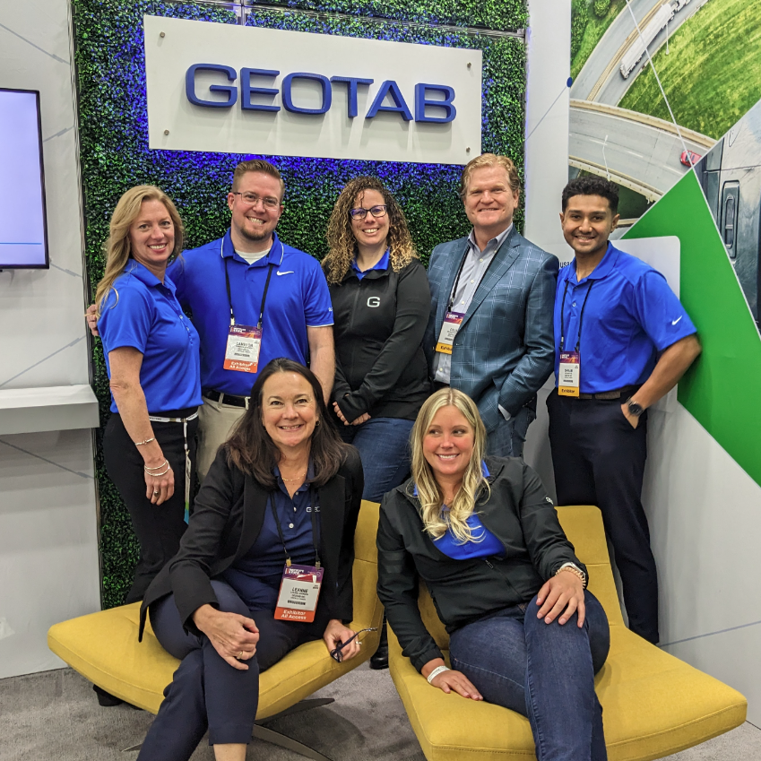 Several geotabbers stand in two rows and smile. Five people are standing in front of the geotab logo, and two girls are sitting on chairs.