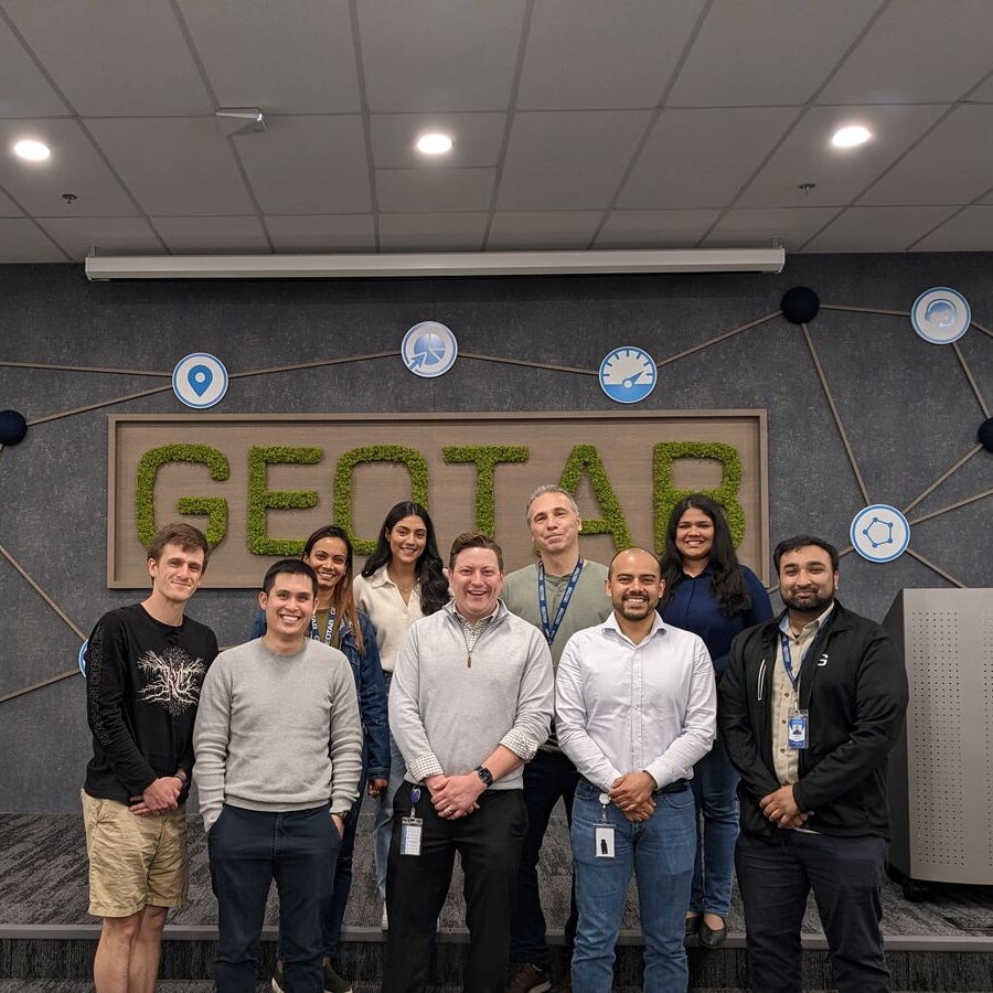 A team of several geotabbers stand in front of a gray wall with a large green geotab logo.