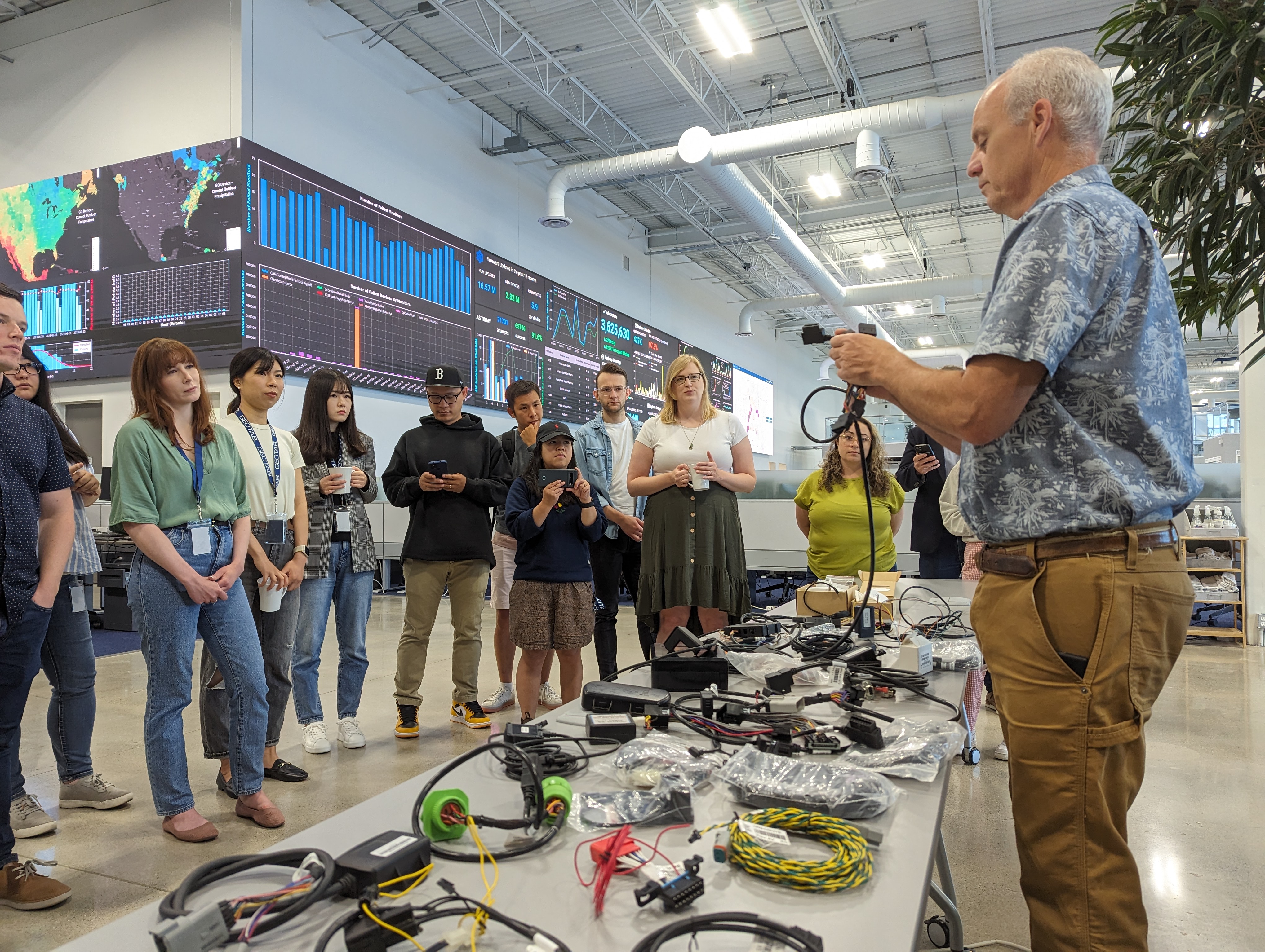 A man demonstrates electronic wiring and components to a diverse group of people in the Geotab office