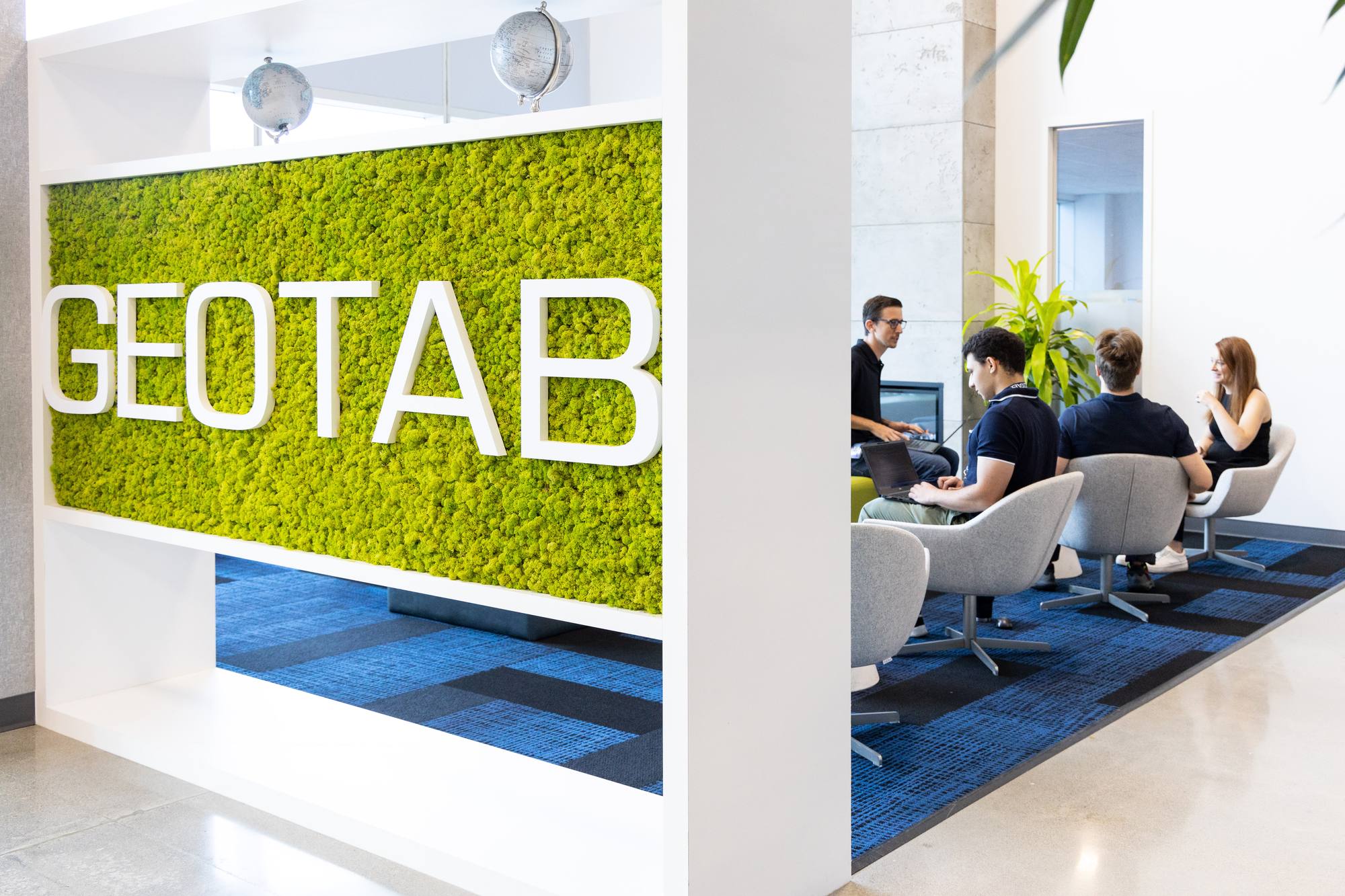 The Geotab logo on a moss wall, with Geotab employees chatting in a lounge area in the background.