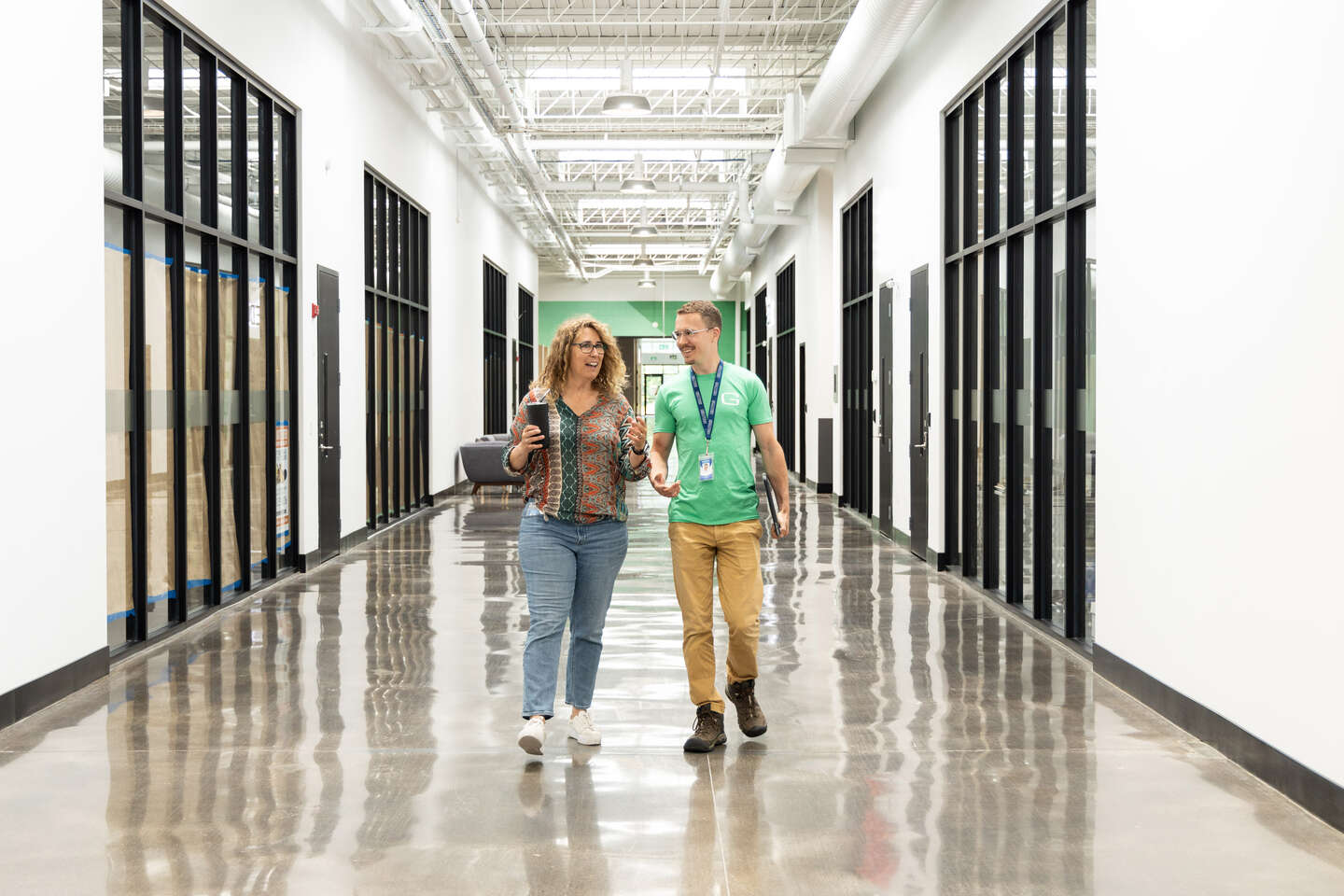 A woman in casual clothes walks along a bright corridor and talks with a guy in a green geotab T-shirt.