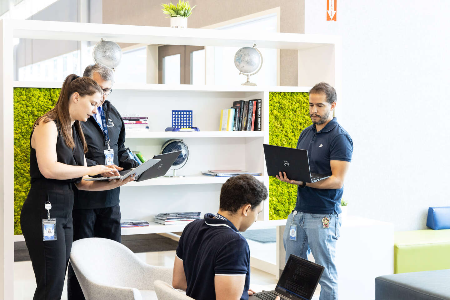 Four people in black T-shirts walk around the bright room and look at laptops.