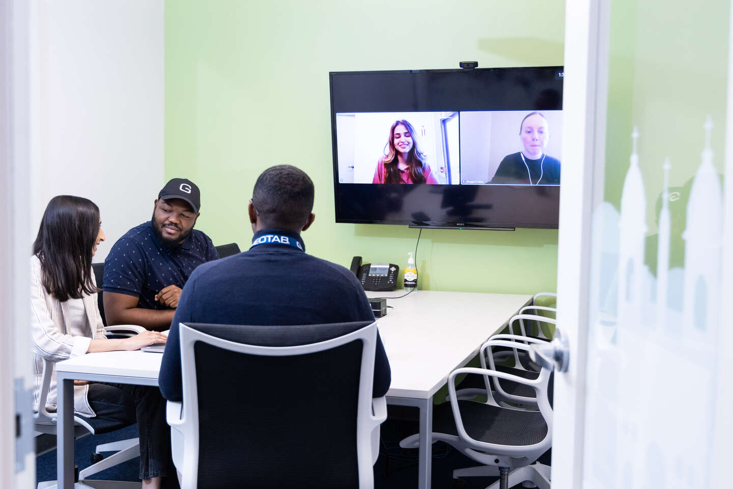 Three geotabbers sit at a white table. On the wall is a screen on which a video conference with two women.