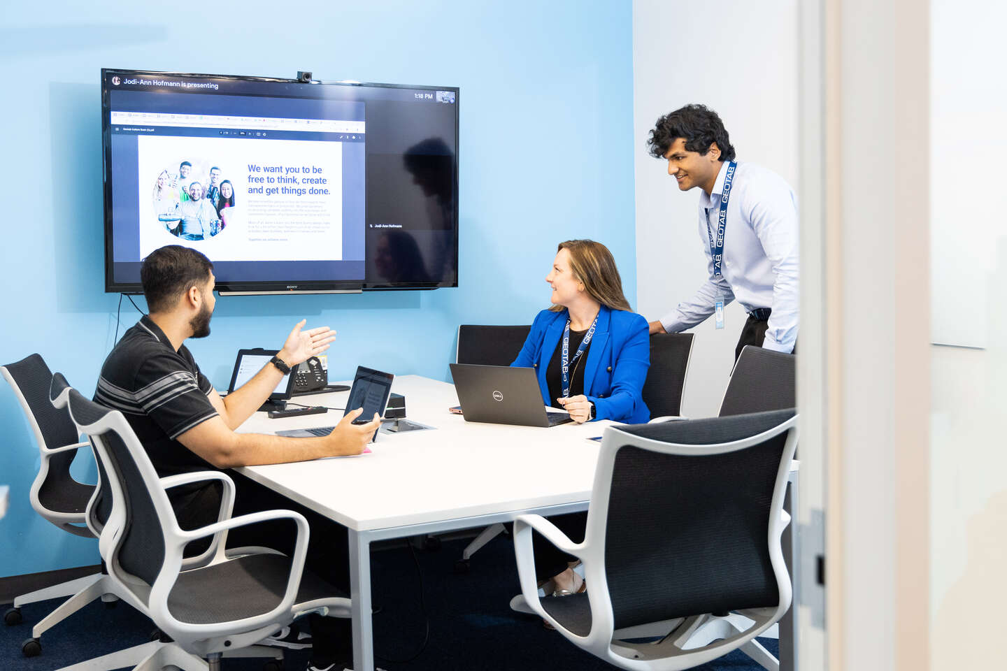 Two geotabbers in the office sit across from a man in a black shirt. A woman in a blue suit is looking at a large information screen on the wall.