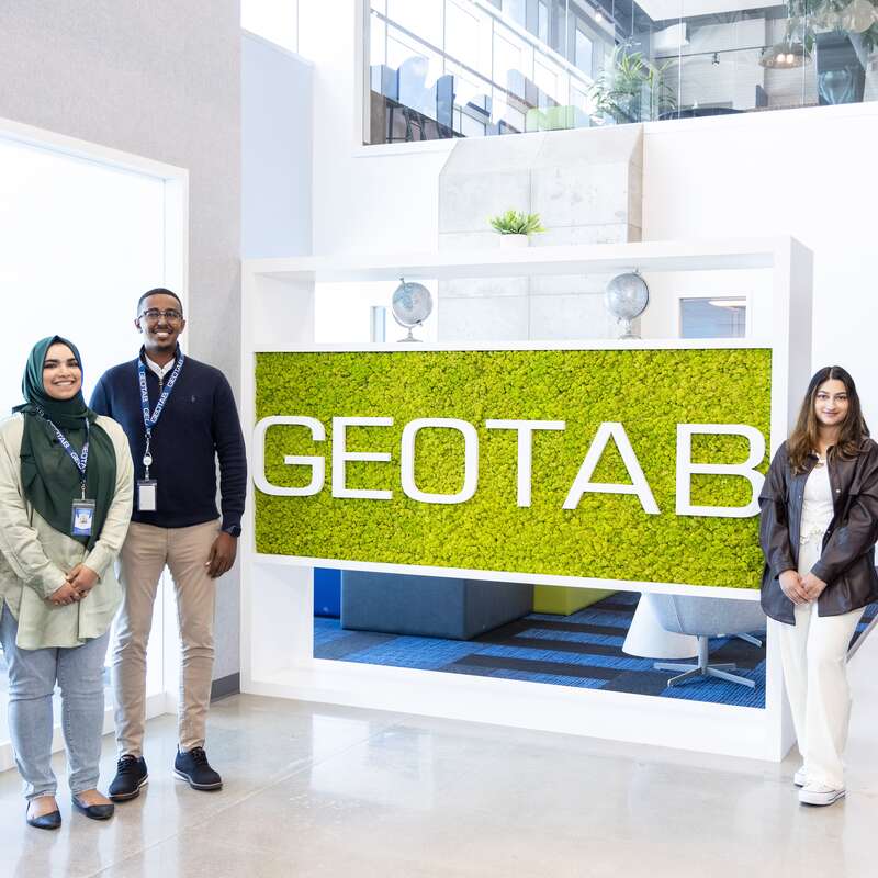 Three Geotab employees stand on different sides of a large stand with the geotab logo on a green background.